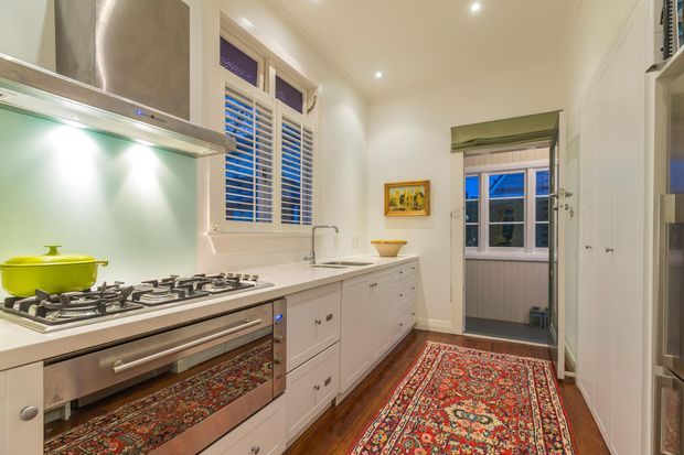 A narrow, traditional-style kitchen with white shaker cabinetry, a gas cooktop and stainless-steel rangehood, timber flooring, patterned runner rugs, plantation shutters, and a compact adjoining utility area visible at the rear. Recessed ceiling lights and warm finishes create a refined, inviting atmosphere.