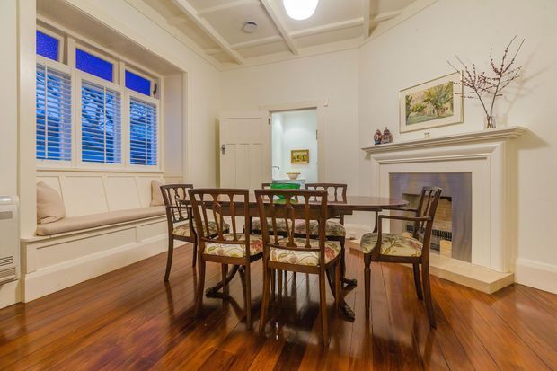 Traditional dining room with a polished dark timber dining table and chairs, a built-in window seat, detailed coffered ceiling, decorative fireplace mantel, framed artwork, and warm timber flooring. White painted mouldings and plantation shutters create a refined, heritage-inspired interior.