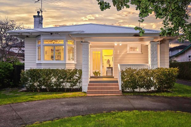 Front exterior view of a single-storey suburban bungalow at dusk, featuring a low-pitched corrugated metal roof, white horizontal weatherboard cladding, enclosed porch, decorative square columns, front steps, and illuminated windows and entry door. Hedges and a paved driveway frame the facade.