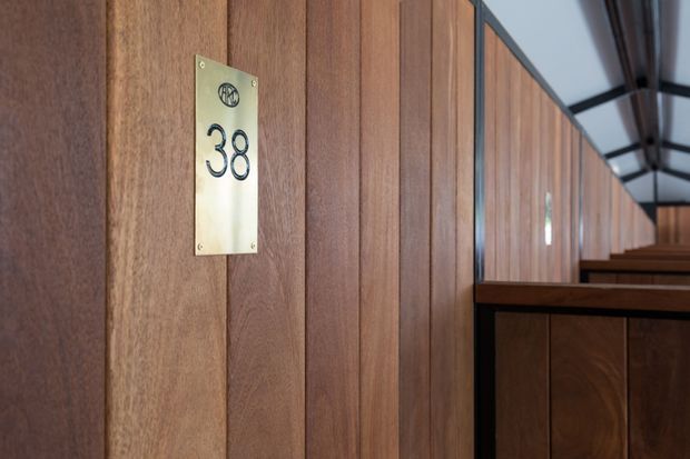 Close-up architectural view of a warm timber-paneled interior corridor or passage, featuring a brass numbered plaque marked 38, dark trim, and repeated timber partitions extending into the distance. The space has a refined, traditional institutional or hospitality character with a partially visible glazed or skylit roof structure.