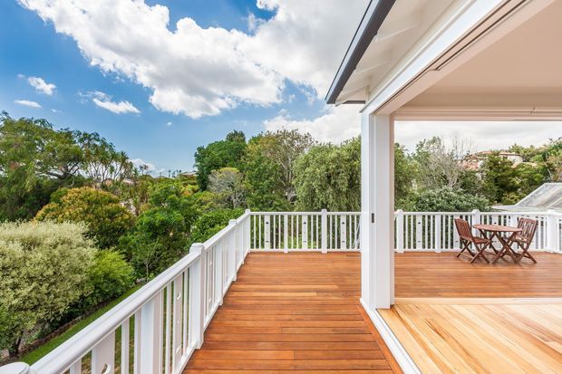 A bright elevated outdoor timber deck and covered terrace with white-painted posts, railings, fascia and ceiling. The deck overlooks dense leafy vegetation and neighbouring homes, with a small outdoor dining table and chairs beneath the covered section. The image has a sunny, airy residential setting with expansive sky and greenery.