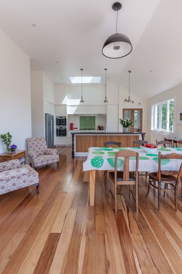Bright open-plan interior combining a dining area, living seating and kitchen. The room features polished warm timber floorboards, white walls and cabinetry, a central kitchen island with wood-panelled detailing, pendant lighting, skylight, large windows and floral upholstered armchairs. A timber dining table with mismatched wooden chairs occupies the foreground.