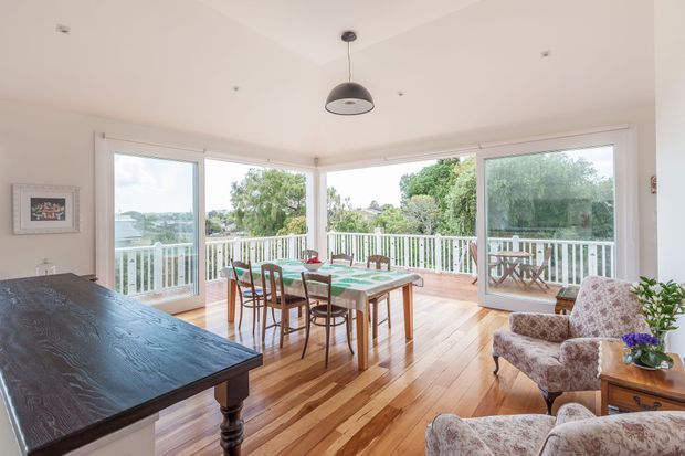 Bright, airy dining and living area with extensive corner glazing opening onto a timber deck and leafy suburban outlook. The room features polished timber flooring, white walls and trim, a dark pendant light, a timber dining table with mismatched chairs, and upholstered armchairs. A large dark timber surface occupies the foreground.