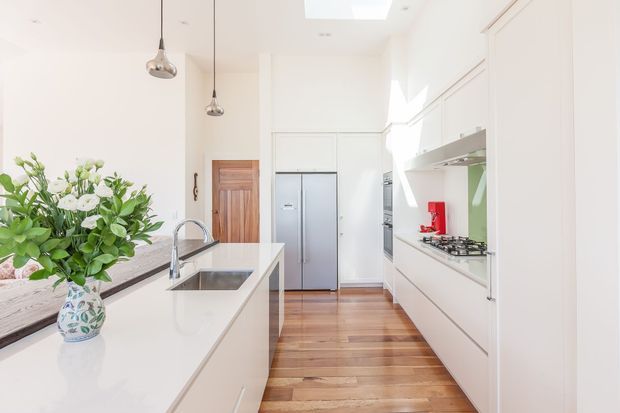 Bright contemporary white kitchen with a long island featuring an inset sink and chrome mixer tap, white flat-panel cabinetry, integrated appliances, a stainless-steel refrigerator, and a pale stone-looking benchtop. Warm timber flooring, a timber internal door, pendant lights, and a skylight add contrast. A large floral arrangement sits on the island, and strong daylight fills the narrow galley-style space.