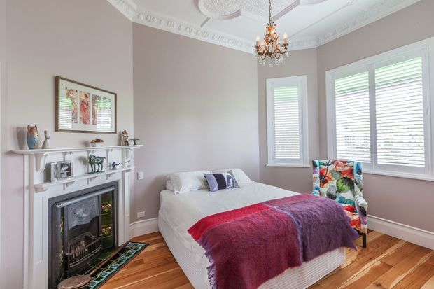 A bright, traditional bedroom with a white upholstered bed, colourful throw and accent pillows, and a vibrant patterned armchair. The room features a decorative white fireplace mantel with a dark insert, pale taupe walls, timber flooring, plantation shutters, ornate ceiling cornices and medallions, and a small crystal chandelier.