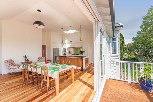Bright open-plan dining and kitchen area opening onto a timber deck or balcony through extensive white-framed glazing. The room features polished timber flooring, a large timber dining table with wooden chairs, a warm timber kitchen island with a dark benchtop, white walls and cabinetry, pendant lighting, and a skylight. Lush greenery is visible outside.