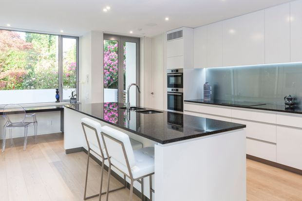 Bright contemporary kitchen with a large central island and seating for two, glossy white full-height cabinetry, integrated stainless-steel appliances, a dark polished stone benchtop, and a pale glass splashback illuminated by under-cabinet lighting. Floor-to-ceiling glazing and a glazed door open to leafy planting outside, while pale timber flooring adds warmth.