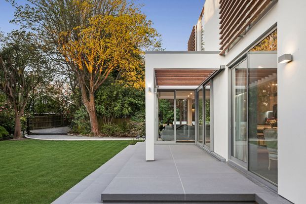 A contemporary residential courtyard or side garden viewed from the outdoor terrace. The scene features a clean white rendered facade, extensive floor-to-ceiling glazing, a broad grey tiled terrace with steps, and timber slatted screening and pergola elements. Mature trees and manicured lawn border the space, while the glazed interior reveals part of a furnished living area.