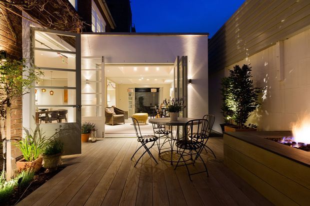 An evening view of a contemporary courtyard-style outdoor entertaining area, with a timber deck, black metal dining furniture, potted plants, and a long built-in outdoor fireplace or fire feature. Large glazed doors open into a brightly lit interior living space, while warm architectural lighting highlights the surrounding modern walls and planting.