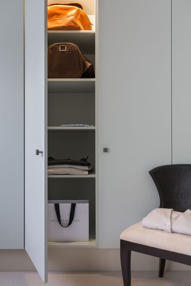 A close-up interior view of a built-in wardrobe or dressing storage unit with pale grey cabinet doors, open shelves containing folded clothing and handbags, and a dark upholstered chair with a light cushion in the foreground. The space is softly lit with a restrained neutral palette and tailored, refined detailing.