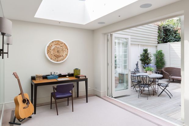 Bright, refined home office or study opening directly onto a furnished outdoor terrace through wide glazed folding doors. The room has pale walls, light carpet, a slim black desk, purple upholstered chair, guitar, decorative round wall artwork, recessed ceiling lights, and a large skylight. Outside is a small timber-decked courtyard terrace with a café table, chairs, and planted screening.