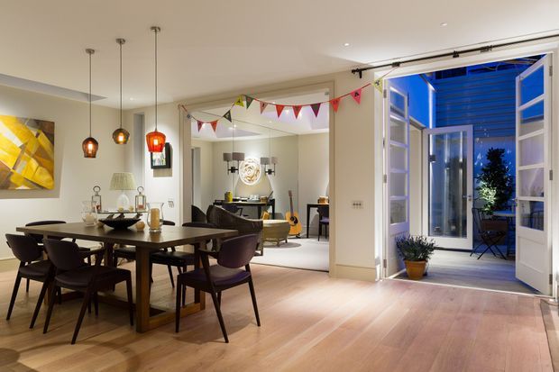 Open-plan dining and living area with a timber dining table and dark upholstered chairs, pendant lights, light timber flooring, and neutral walls. Festive bunting spans the room, while large bi-fold doors open onto a small outdoor terrace or balcony at dusk. A living room and console area are visible beyond the dining space.