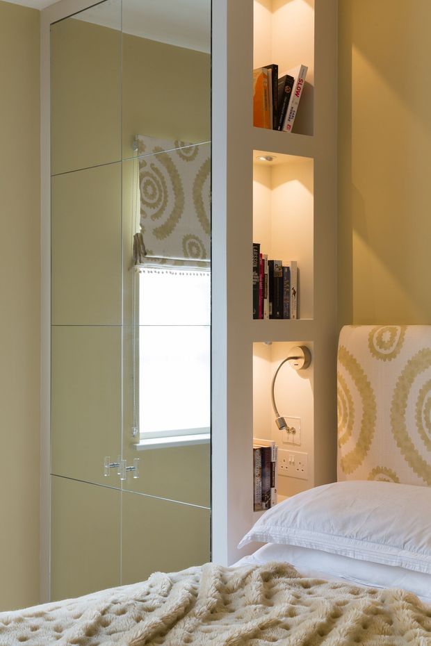 A warm, compact bedroom detail featuring a bed with a patterned upholstered headboard, white bedding, and a textured cream throw. Beside the bed is a built-in recessed shelving unit with integrated warm lighting, books, and a small reading lamp. Mirrored wardrobe or wall panels reflect the window and headboard, making the room feel brighter and more spacious.