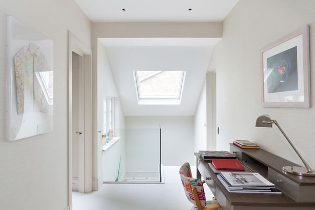 Bright, narrow upstairs workspace or study area with a built-in desk and shelving along the right wall, a desk lamp, books and papers, framed artwork, and a skylight above a stairwell or landing. The space has white and pale neutral finishes, abundant natural light, and a clean contemporary feel.