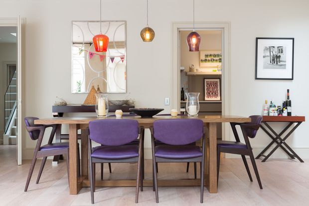 A refined dining room with a large timber dining table surrounded by purple upholstered mid-century-style chairs. Three pendant lights hang above the table, while a mirrored artwork, sideboard, framed photograph, and drinks console occupy the pale neutral walls. An open doorway reveals a small pantry or kitchen storage area beyond.