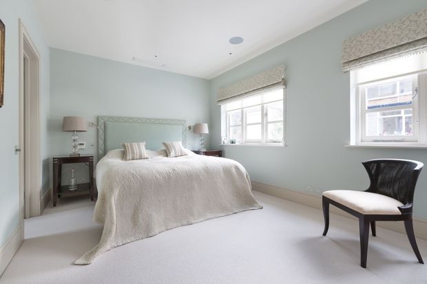 Bright, serene bedroom with pale blue-green walls, a double bed dressed in a white textured coverlet, upholstered geometric headboard, dark bedside tables, matching lamps, white-framed windows with patterned Roman blinds, and a simple dark accent chair. The room has a soft neutral carpet, white ceiling and trim, and a calm traditional/contemporary feel.