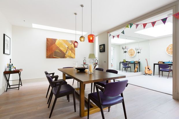 Bright contemporary dining room with a large timber dining table and purple upholstered dining chairs. The room features pale timber flooring, white walls and ceiling, colourful pendant lights, abstract wall art, and a wide glazed opening into an adjacent light-filled sitting or studio area with additional seating and a guitar. Decorative bunting adds a playful accent.
