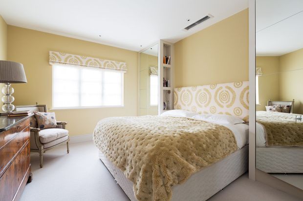 A bright, warmly styled bedroom with pale yellow walls, a large upholstered bed, cream textured bedding, and a patterned upholstered headboard wall panel. A mirrored wardrobe reflects the bed, while a built-in open bookshelf, armchair, timber dresser, table lamp, and softly filtered window light complete the room.