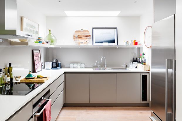 Bright contemporary galley-style kitchen with white walls and ceiling, pale timber flooring, white benchtops, streamlined taupe-grey handleless cabinetry, stainless steel appliances, and open shelving displaying kitchenware. A skylight and recessed ceiling lights provide abundant natural and artificial light.