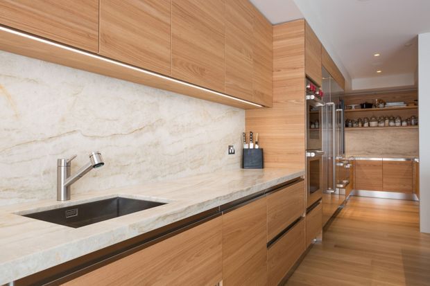 A refined contemporary galley kitchen with extensive warm timber cabinetry, a pale veined stone benchtop and matching splashback, integrated stainless-steel appliances, under-cabinet lighting, and a timber floor. The long linear layout leads toward additional cabinetry and open shelving at the rear.