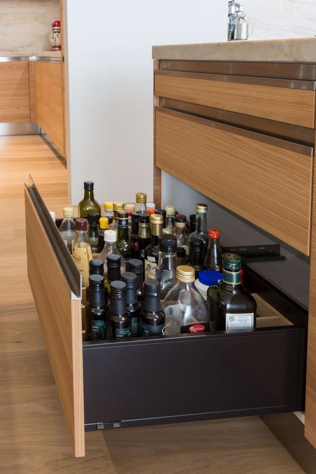 Close-up view of a contemporary kitchen featuring a large open timber-fronted drawer filled with bottles and pantry items. The cabinetry has warm natural wood grain, dark internal drawer boxes and slim metallic edge details, with a light stone benchtop and matching timber flooring visible in the background.