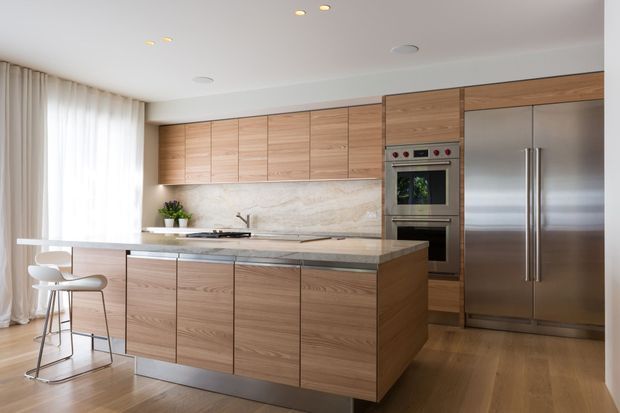 A bright, contemporary kitchen with extensive light timber cabinetry, a large central island with seating, pale stone benchtops and splashback, stainless steel integrated appliances, and floor-to-ceiling sheer curtains. The space has timber flooring, recessed lighting, and a clean, refined open-plan feel.