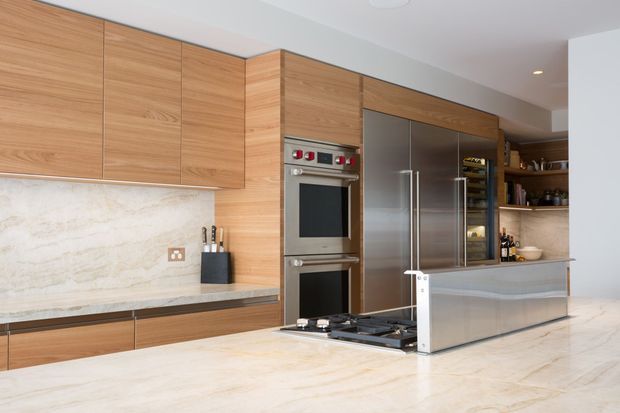 Contemporary kitchen with extensive warm timber cabinetry, integrated double ovens, a large stainless-steel refrigerator, stone benchtops and a pale veined stone splashback. A central cooking area with a glass-and-metal guard sits behind a large island or peninsula, with open shelving and pantry storage at the rear.