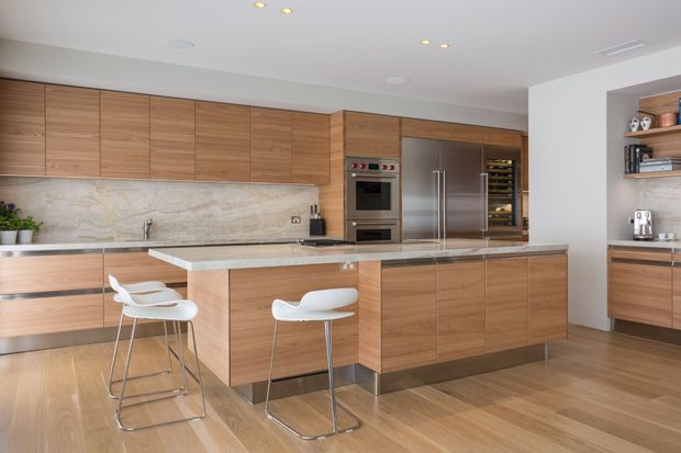 Bright, spacious contemporary kitchen with extensive warm timber cabinetry, a large central island with pale stone benchtop, integrated stainless-steel appliances, and a natural stone-look splashback. Two white bar stools sit at the island, while open shelving and additional joinery line the right wall. Timber flooring and recessed ceiling lighting create a clean, refined finish.