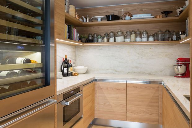 Compact, well-organised contemporary kitchen with warm timber cabinetry, pale stone benchtops and a matching full-height stone splashback. Open shelving displays crockery, cookbooks and glass storage jars, while integrated stainless-steel appliances include a wine refrigerator and oven. A red stand mixer provides a strong accent colour.
