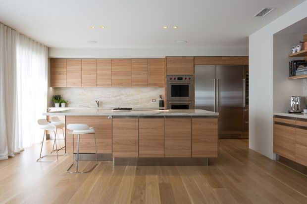 A contemporary open-plan kitchen with extensive warm timber cabinetry, a large central island with pale stone benchtop, integrated stainless-steel appliances, and a light stone backsplash. Floor-to-ceiling sheer curtains and timber flooring create a bright, refined interior, with a small dining or breakfast area and built-in shelving visible at the right.