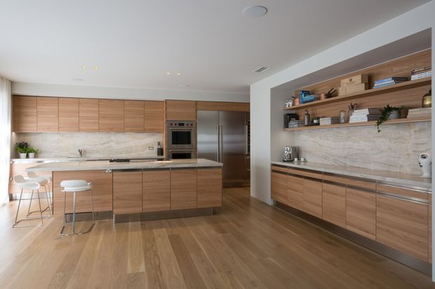 Contemporary open-plan kitchen with extensive warm timber cabinetry, a large central island with bar seating, integrated stainless-steel appliances, and a pale marble-look splashback. Open timber shelves with decorative objects and greenery line the right wall, while wide timber flooring and recessed ceiling lighting create a clean, refined feel.