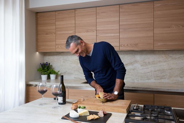 A warm, contemporary kitchen with extensive light timber cabinetry, a pale stone-look splashback, and a large island bench. A man is preparing food on a timber chopping board beside a gas cooktop, with wine glasses and ingredients on the island.