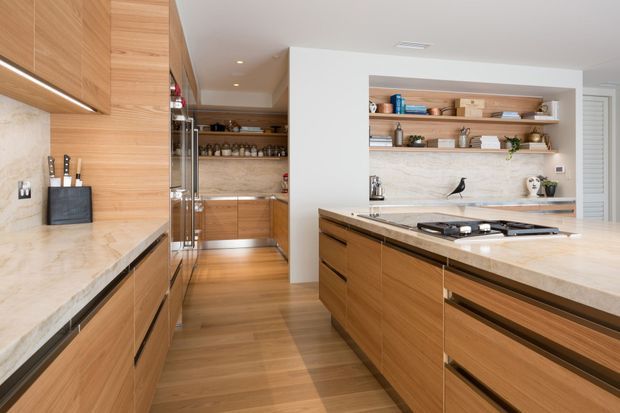 Warm contemporary kitchen with extensive light timber cabinetry, a large central island with integrated gas cooktop, pale stone benchtops, open display shelving, and timber flooring. A secondary preparation or pantry area is visible through the opening at the rear.