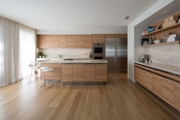 A bright, contemporary open-plan kitchen with extensive warm timber cabinetry, a central island with white stools, integrated stainless-steel appliances, and a pale veined stone backsplash and benchtop. Full-height sheer curtains and pale timber flooring create an airy, refined atmosphere, while open shelving and a secondary run of cabinetry line the right wall.