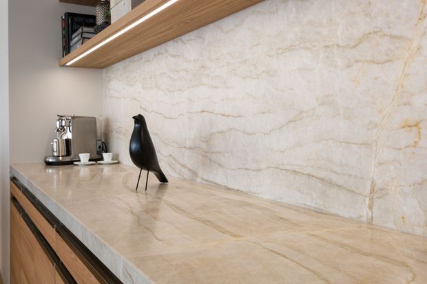 Close-up of a refined kitchen or beverage station featuring a large cream-and-beige veined stone benchtop and matching full-height splashback. Warm timber cabinetry and an open timber shelf with integrated linear lighting add contrast, while a coffee machine and sculptural black vase sit on the counter.