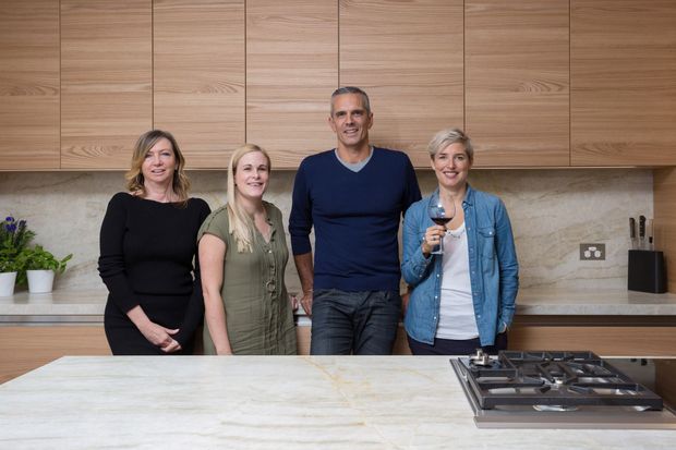 A contemporary kitchen photographed from the island, featuring full-height light timber cabinetry with vertical panel divisions, a pale stone or marble-look splashback and benchtop, integrated under-cabinet lighting, and a dark built-in gas cooktop. Four people are standing in the kitchen, with small plants and a knife block on the rear benchtop.