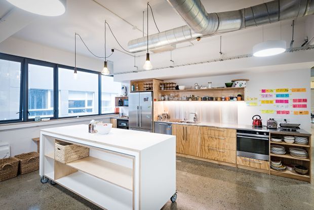 Bright open-plan commercial-style kitchen or staff breakout space with a large white mobile island, extensive timber cabinetry, stainless-steel appliances and splashback, exposed silver ventilation ducts, pendant lights, and large black-framed windows. Colourful notes are arranged on the wall, and open shelving displays crockery and kitchenware.