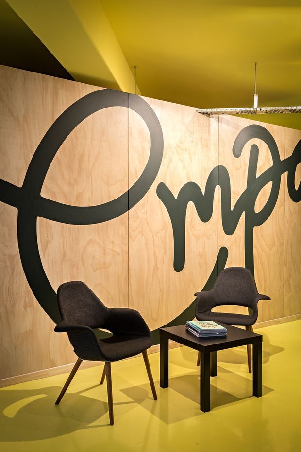 A boldly styled commercial interior or exhibition-style lounge with a vivid mustard-yellow ceiling and floor, pale timber plywood wall panels, and oversized black graphic lettering across the walls. Two dark upholstered lounge chairs and a small black square coffee table with magazines form a compact seating area. Suspended track lighting and exposed ceiling services contribute to the graphic, industrial atmosphere.