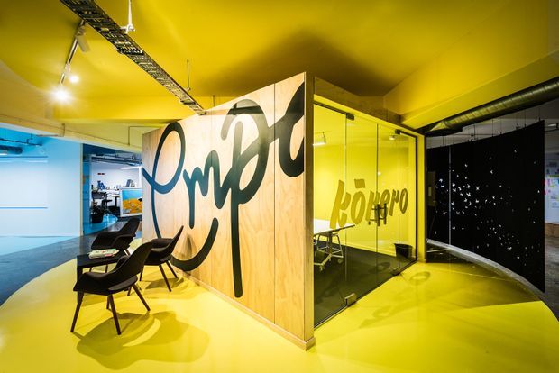 A vivid contemporary commercial interior with a bright yellow floor and ceiling, a glass-enclosed meeting room, timber-panelled partitions featuring oversized black graffiti-style lettering, exposed black ductwork, track lighting, and lounge chairs. The space has an open-plan, creative studio or office character with bold graphic branding and high visual contrast.