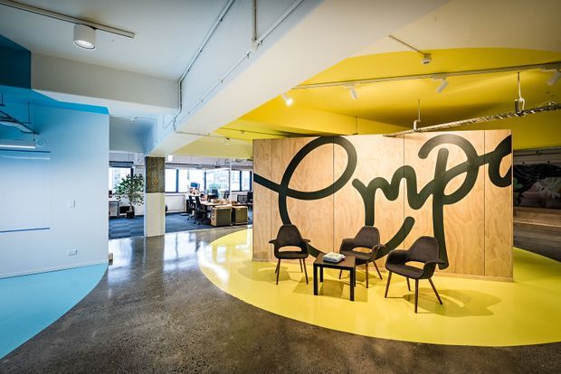 A vibrant contemporary commercial interior with a polished concrete floor, curved yellow floor and ceiling zones, bright cyan walls, exposed ceiling services, track lighting, and a large plywood feature partition with bold black lettering. A small seating arrangement sits in front of the partition, while an open office workspace is visible beyond.
