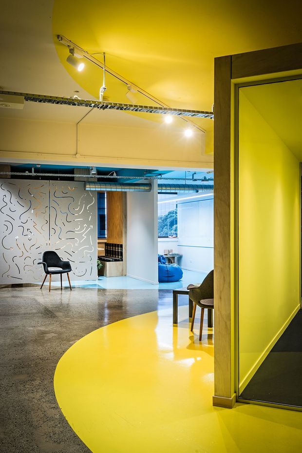 A vibrant contemporary commercial interior with an open-plan workspace or studio. The space features a polished concrete floor intersected by a bold glossy yellow circular floor zone, yellow walls and ceiling, exposed services and ductwork, track lighting, timber-framed glazing, and a decorative perforated white partition with abstract line patterns. Lounge and work seating are visible deeper in the space, with strong blue accents and large windows.