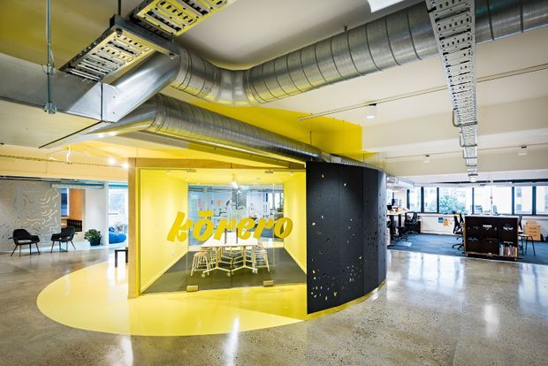 A bright contemporary open-plan commercial office interior with polished concrete flooring, exposed silver HVAC ductwork and cable trays, and a bold yellow glazed meeting room or collaboration pod at the centre. The space includes shared desks, office workstations, chairs, and large windows, with a black perforated feature wall beside the yellow enclosure.