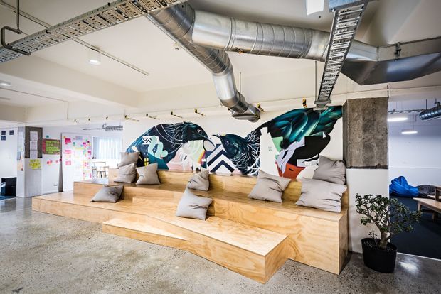 An open-plan commercial interior with tiered plywood bleacher-style seating, scattered cushions, colourful graphic wall art, exposed concrete columns, polished concrete flooring, and visible exposed ductwork and cable trays across the ceiling. The space has a bright, informal creative-office atmosphere.