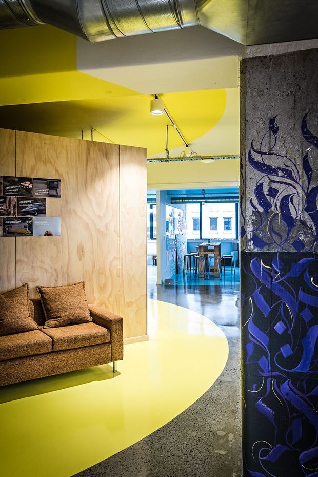 A vibrant contemporary interior with a yellow painted floor and ceiling featuring large curved graphic shapes. A brown upholstered sofa sits beside a plywood partition wall with displayed prints. Exposed silver HVAC ductwork, track lighting, polished concrete flooring, and a blue-and-purple patterned structural column contribute to an eclectic industrial character. Through the opening, a compact dining or breakout area with a high table and stools is visible.