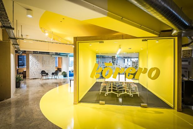 A vibrant contemporary commercial office interior with a bright yellow glazed meeting room or collaboration pod at the centre. The pod has full-height glass doors and walls, yellow branding lettering, a conference table with rolling chairs, and a polished concrete floor with a large curved yellow floor and ceiling treatment. Exposed services, including ductwork and cable trays, contribute an industrial office character. Additional open workspaces and seating are visible in the background.