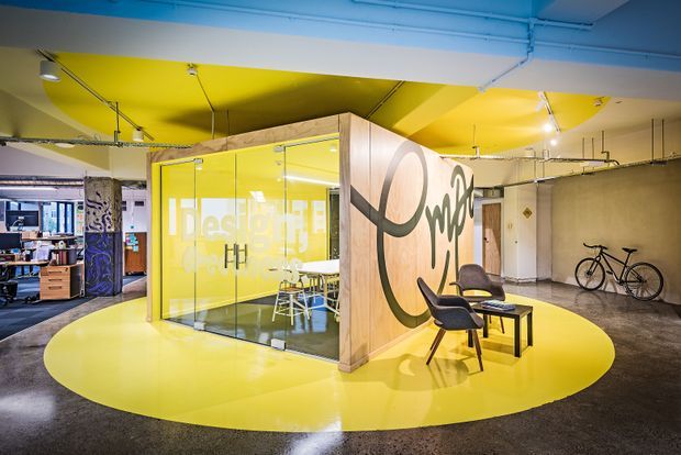 A bright, open-plan creative office interior with a glazed yellow meeting room at the centre, graphic lettering on timber-clad walls, exposed services and lighting tracks, and a vivid circular yellow floor feature. Lounge chairs and a small coffee table form a seating area in the foreground, with workstations visible beyond. The space has an industrial shell enlivened by bold contemporary branding and colour.