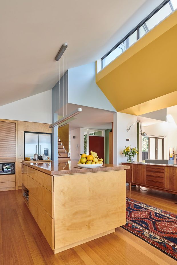 A bright, open-plan kitchen with a large central timber island, integrated shelving and cabinetry, and a separate timber sideboard. The space features warm timber flooring and joinery, white walls, a dramatic mustard-yellow ceiling and bulkhead, clerestory windows, exposed vertical circulation elements, and a colourful patterned rug. A dining or living area and staircase are visible beyond the kitchen.