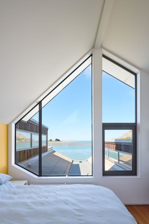 A bright bedroom with a partially visible bed facing dramatic angular floor-to-ceiling corner glazing. White walls and ceiling, dark window frames, and timber flooring surround expansive views of turquoise water, a sandy shoreline, and nearby rocky landforms. The glazing forms an architectural triangular composition beneath a sloping ceiling, with an exterior deck and neighbouring timber-clad structure visible outside.