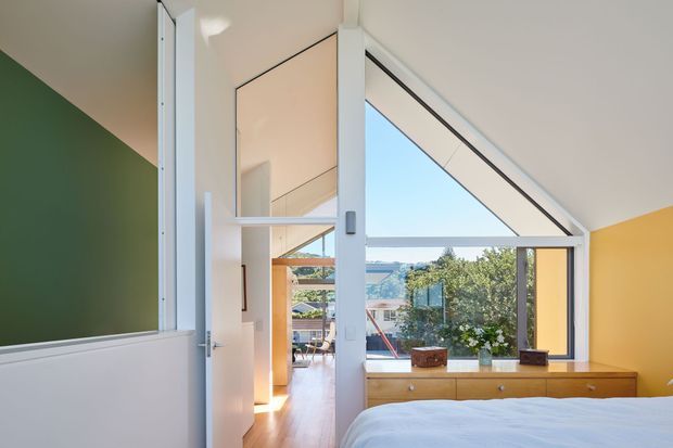 Bright contemporary bedroom viewed toward a dramatic triangular glazed gable window, with a partial view of an adjoining hallway or living area through an open white door. The room has white walls and ceiling, a deep green accent wall on the left, a warm yellow-orange wall at right, timber flooring, and a long built-in timber dresser beneath the window. The glazing provides views toward neighbouring rooftops and greenery, while the angled ceiling and window create a distinctive architectural feature.