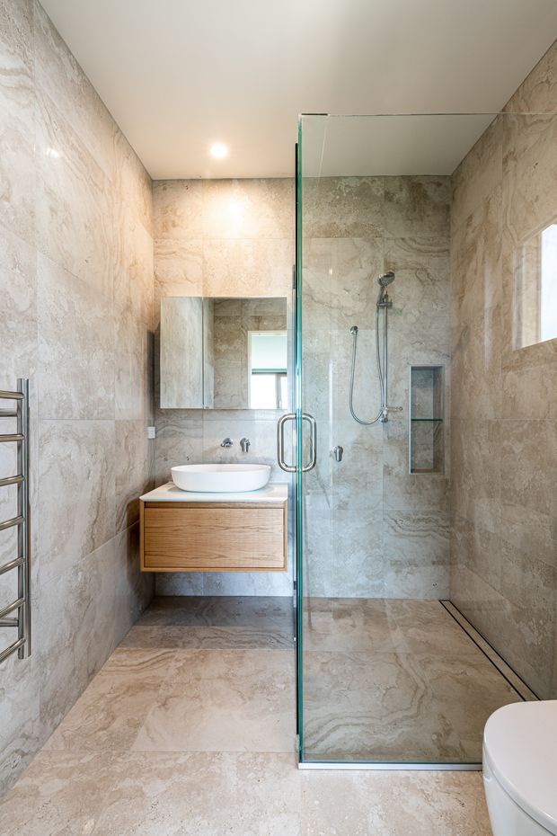 A contemporary bathroom featuring full-height beige natural-stone-look wall and floor tiles, a frameless glass walk-in shower with dual shower fixtures and recessed niche, a floating timber vanity with a white vessel basin, mirrored cabinet, chrome heated towel rail, and partially visible toilet. The room is compact, bright, and refined with a cohesive neutral palette.